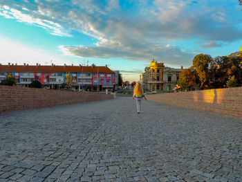 Rear view of person walking on footpath by buildings against sky