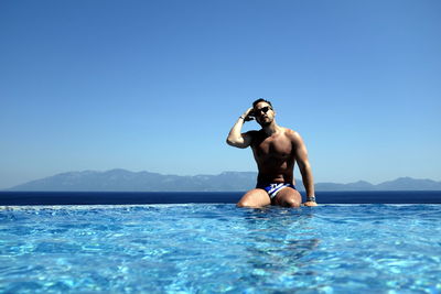 Young man sitting at infinity pool against clear sky on sunny day