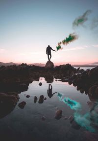 Man standing on rock by lake against sky