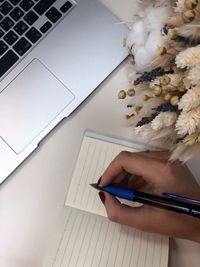 Cropped hand of woman writing in book by laptop on table