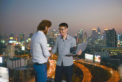 Colleagues discussing over laptop while standing on building terrace against illuminated city at night