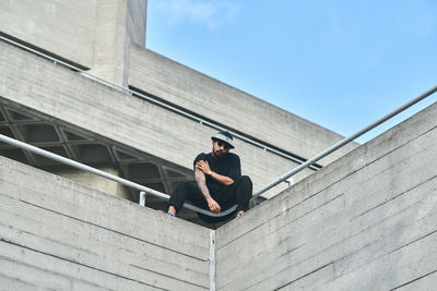 Low angle view of young man wearing sunglasses while sitting on building against blue sky