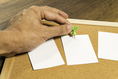 Cropped hand of man holding thumbtack with paper on bulletin board