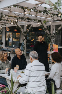 Smiling male waiter serving food to senior customers sitting at restaurant