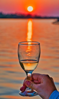Midsection of person holding wineglass against orange sky during sunset