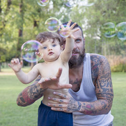 Smiling bearded man holding son playing with bubbles in park