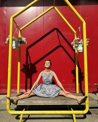 Beautiful woman sitting with legs apart amidst hanging potted plants on bench against red metallic wall