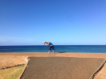Rear view of woman exercising by beach against clear blue sky