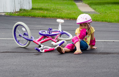 A little girl in safety gear, sits in the middle of a drive after falling off her new bike.