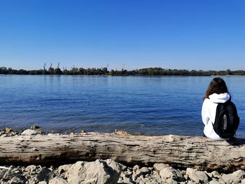 Rear view of a beautiful girl sitting by river against clear sky