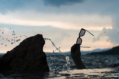 Silhouette man holding rock in sea against sky during sunset