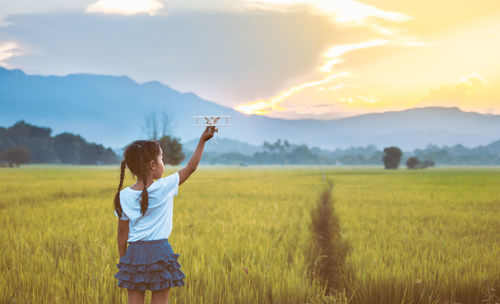 Rear view of girl holding toy airplane on grassy field during sunset