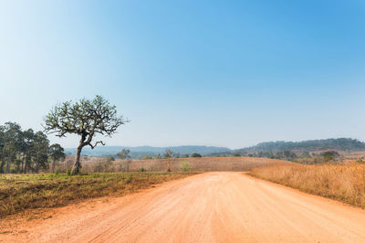 Dirt road by field against clear blue sky