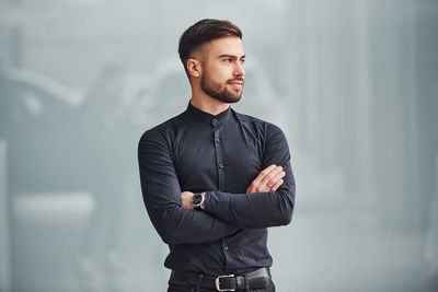 Young bearded guy in elegant clothes standing indoors against grey background.