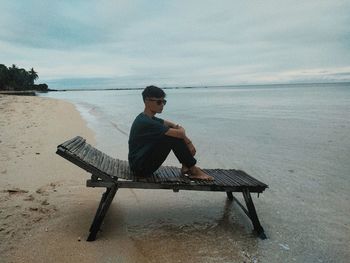 Man sitting on bench at beach against sky