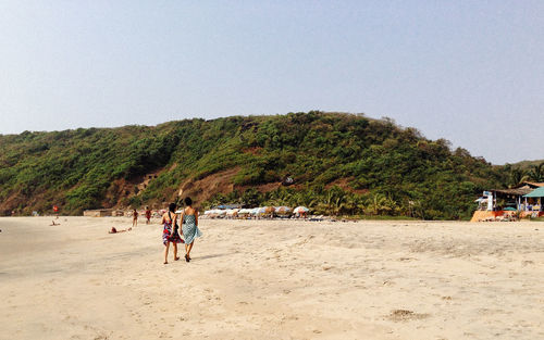 Rear view of women walking at arambol beach by hill against clear sky