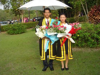 Portrait of smiling people holding bouquets wearing graduation gown standing at park