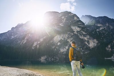 Man with camera standing by lake against mountain during sunny day