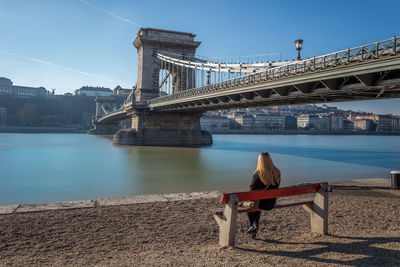 Woman sitting on bench looking at chain bridge over river danube in city