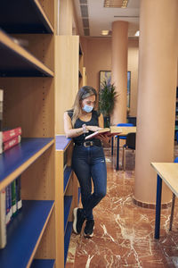 A young girl with a mask is standing in the library looking at a book