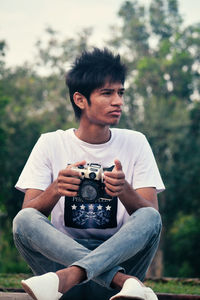 Young man sitting with camera against trees