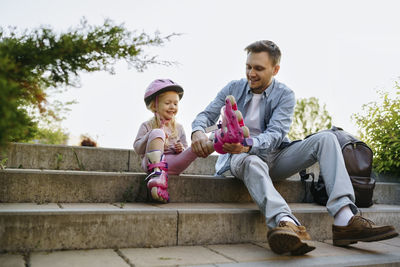 Smiling man helping daughter to wear roller skates sitting on steps