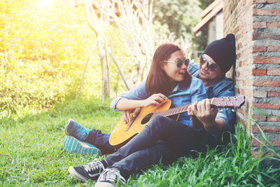 Happy couple looking at each other while playing guitar while sitting on grassy field against brick wall