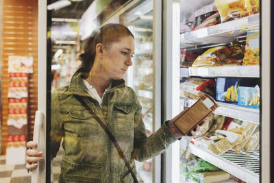 Female customer reading product seen through glass door at refrigerated section