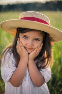 Village girl child in a white dress and hat stands on a green field at sunset in summer