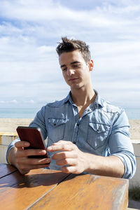 Young man using phone while sitting at cafe against beach