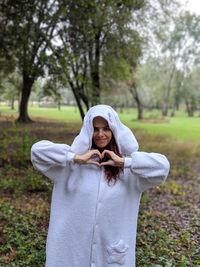 Portrait of young woman wearing white warm clothing while making heart shape against trees
