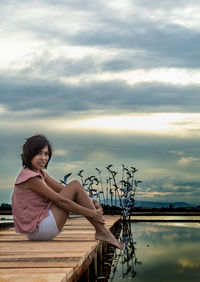 Portrait of mid adult woman sitting on pier over lake against cloudy sky during sunset