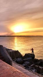 Silhouette man sitting on rock by sea against sky during sunset