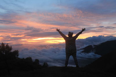 Rear view of man with arms outstretched standing on cliff against sky during sunset