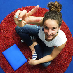 High angle portrait of woman with laptop gesturing peace sign sitting on carpet