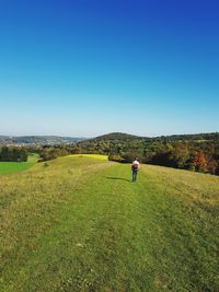 Rear view of woman walking on grassy landscape against clear blue sky