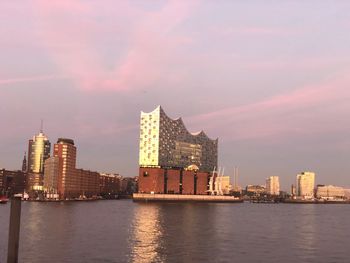 Modern buildings by river against sky at sunset