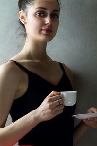 Portrait of confident woman holding tea cup while standing against gray wall
