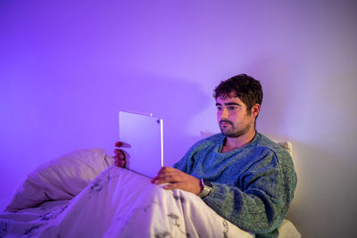 Man using digital tablet sitting on bed against wall at home