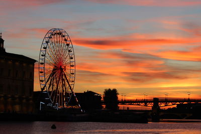 Silhouette ferris wheel by river against sky during sunset