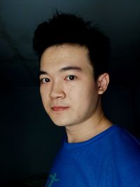 A portrait of a young man wearing a casual blue shirt in his room with black background