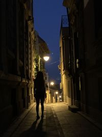 Rear view of woman walking on illuminated street amidst buildings at night