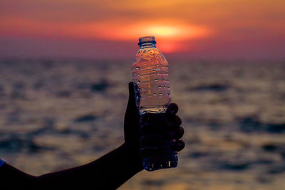 Silhouette man hand holding water bottle by sea against sky during sunset