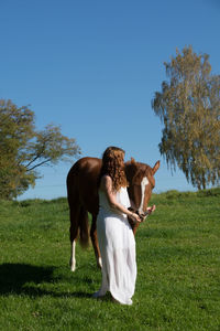 Side view of young woman with horse walking on grassy field against clear sky
