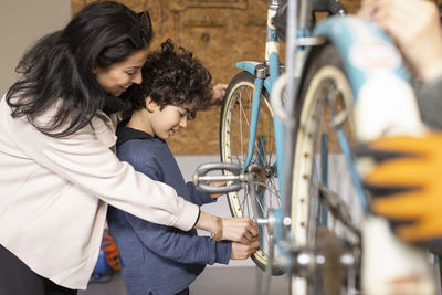 Side view of happy woman assisting son learning to repair bicycle at recycling center