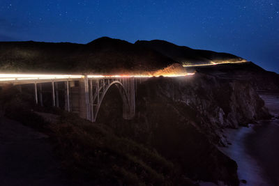 Illuminated bridge over mountains against sky at night