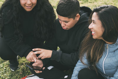 Three people of hispanic-latino ethnicity, sitting on the ground in the park with smart devices