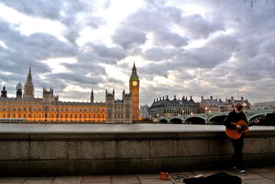 Young man playing guitar while standing against in city cloudy sky