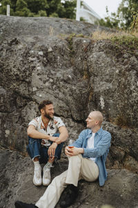 Young male friends having wine and talking to each other while sitting on rocks