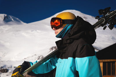 Skier holds a pair of skis and looks at the snow-capped mountains to the side. a guy with skis on